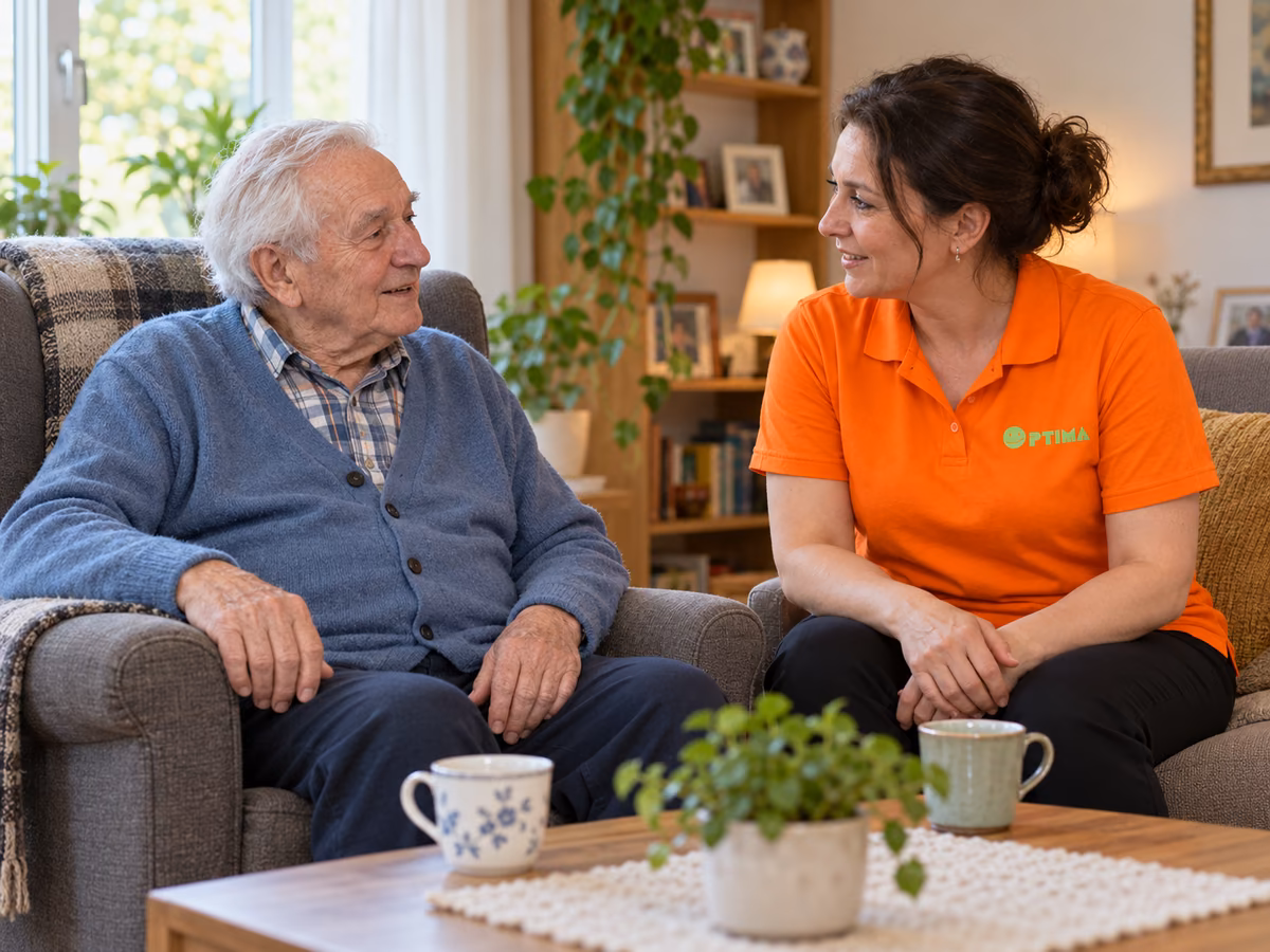Pflegekraft von OPTIMA sitzt bei einem älteren Herrn im Wohnzimmer und unterhält sich in Ruhe mit ihm